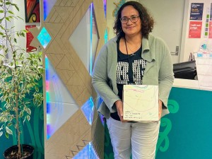 Dr Ramona Tiatia holding a GBB cake box and leaning against a decorated pillar to her left, wiht a potplant on the other side of the pillar.